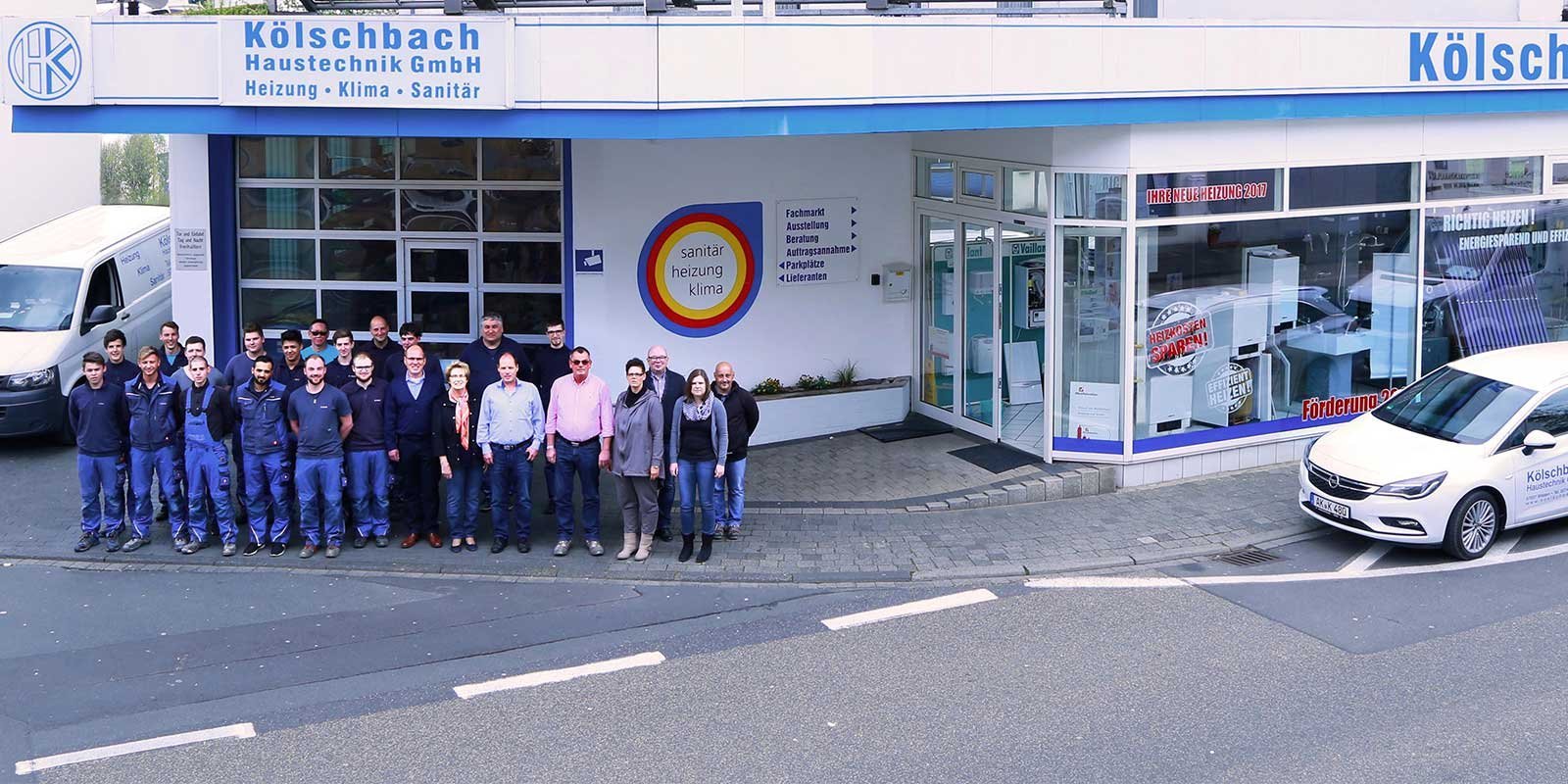 Eine Gruppe von Mitarbeitern steht vor dem Gebäude der Kölschbach Haustechnik GmbH, umgeben von Fahrzeugen und Schildern.